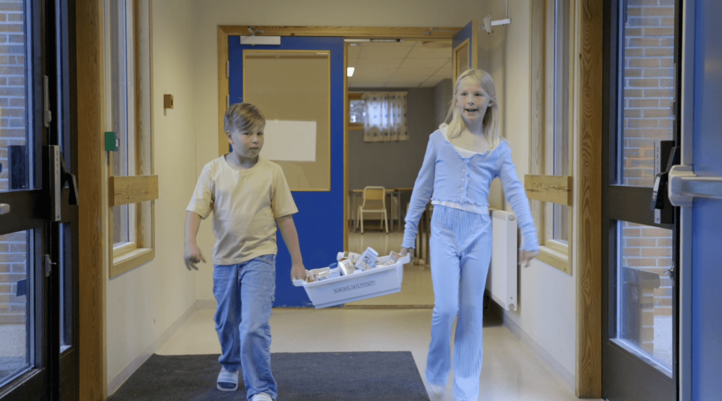 To elever går gjennom en skolekorridor og bærer en kasse med melkekartonger, på vei inn i et klasserom.