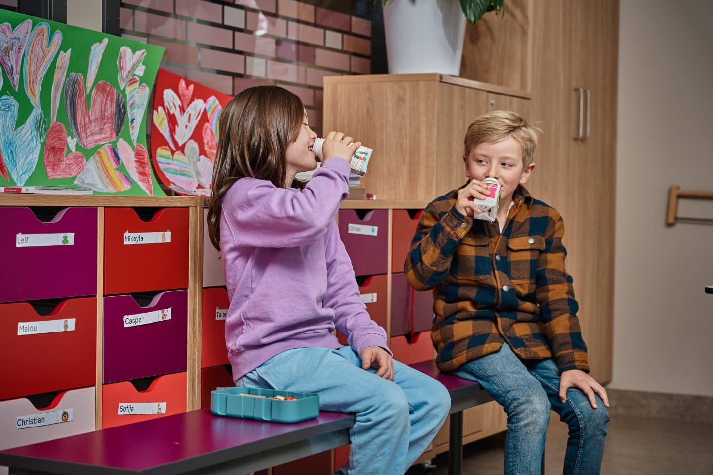 To skolebarn sitter på en benk i klasserommet og drikker skolemelk.